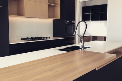 Inspiration for a mid-sized modern galley eat-in kitchen in Christchurch with a double-bowl sink, black cabinets, quartz benchtops, white splashback, glass sheet splashback, black appliances, light hardwood floors, with island and white benchtop.