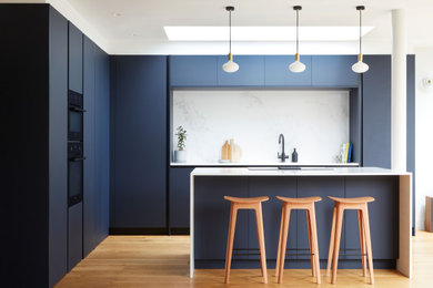 Inspiration for a large contemporary l-shaped open plan kitchen in London with a submerged sink, flat-panel cabinets, blue cabinets, quartz worktops, white splashback, engineered quartz splashback, black appliances, medium hardwood flooring, an island, white worktops and feature lighting.