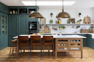 Large traditional l-shaped kitchen/diner in London with a belfast sink, shaker cabinets, green cabinets, quartz worktops, white splashback, engineered quartz splashback, integrated appliances, medium hardwood flooring, an island and white worktops.