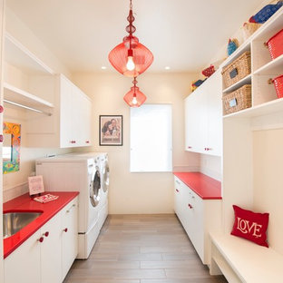 アルバカーキの大きいコンテンポラリースタイルのおしゃれな家事室 (アンダーカウンターシンク、フラットパネル扉のキャビネット、白いキャビネット、ベージュの壁、磁器タイルの床、左右配置の洗濯機・乾燥機、茶色い床、赤いキッチンカウンター) の写真