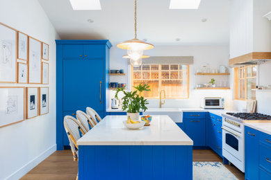 Example of a beach style l-shaped brown floor and medium tone wood floor eat-in kitchen design in San Francisco with a farmhouse sink, blue cabinets, marble countertops, white backsplash, an island, white countertops, shaker cabinets and white appliances