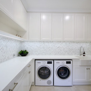 Inspiration pour une grand buanderie en L multi-usage avec un évier de ferme, un placard avec porte à panneau encastré, des portes de placard blanches, un plan de travail en quartz modifié, une crédence grise, une crédence en marbre, un mur gris, un sol en carrelage de céramique, des machines côte à côte, un sol gris et un plan de travail blanc.