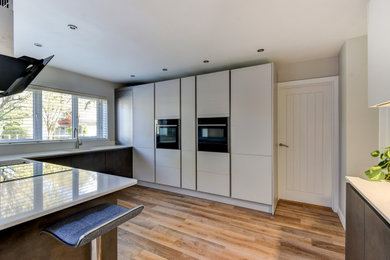 This is an example of a medium sized contemporary grey and white u-shaped kitchen/diner in Sussex with a submerged sink, flat-panel cabinets, grey cabinets, quartz worktops, grey splashback, glass sheet splashback, black appliances, vinyl flooring, an island, brown floors, white worktops and feature lighting.