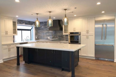 Example of a large transitional l-shaped light wood floor and brown floor enclosed kitchen design in New York with a farmhouse sink, shaker cabinets, white cabinets, marble countertops, brown backsplash, wood backsplash, stainless steel appliances and an island