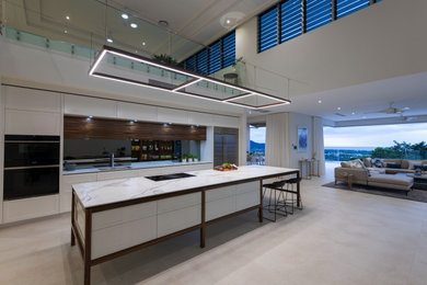 This is an example of an expansive contemporary galley open plan kitchen in Cairns with an undermount sink, recessed-panel cabinets, white cabinets, quartz benchtops, white splashback, mirror splashback, black appliances, porcelain floors, with island, white floor, white benchtop and vaulted.