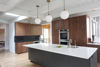 Design ideas for a large midcentury l-shaped open plan kitchen in Raleigh with an undermount sink, flat-panel cabinets, medium wood cabinets, quartz benchtops, black splashback, stone slab splashback, panelled appliances, light hardwood floors, with island, brown floor and white benchtop.