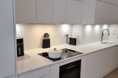 Photo of a large modern single-wall open plan kitchen in London with a built-in sink, flat-panel cabinets, white cabinets, quartz worktops, white splashback, stone slab splashback, white appliances, painted wood flooring and white worktops.