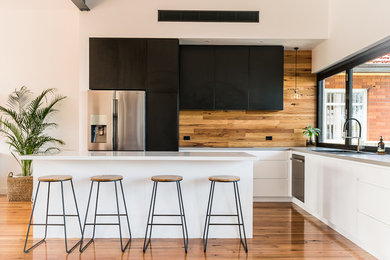 This is an example of a mid-sized l-shaped open plan kitchen in Wollongong with an undermount sink, black cabinets, quartz benchtops, timber splashback, black appliances, medium hardwood floors and with island.