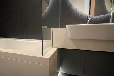 This is an example of a medium sized contemporary grey and brown family bathroom in London with flat-panel cabinets, brown cabinets, a built-in bath, a shower/bath combination, a wall mounted toilet, brown tiles, stone slabs, brown walls, marble flooring, a built-in sink, quartz worktops, brown floors, a hinged door, white worktops, feature lighting, a single sink and a built in vanity unit.