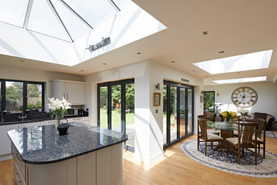 Large contemporary l-shaped open plan kitchen in Berkshire with white cabinets, granite worktops, black splashback, granite splashback, light hardwood flooring, an island and black worktops.