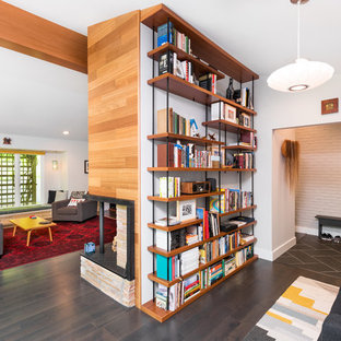 This is an example of a large midcentury home office in Detroit with white walls, dark hardwood floors, a brick fireplace surround, brown floor, a library and a two-sided fireplace.