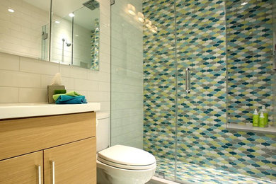 Photo of a mid-sized transitional 3/4 bathroom in New York with flat-panel cabinets, light wood cabinets, an alcove shower, multi-coloured tile, white walls, an integrated sink, a one-piece toilet, ceramic tile, ceramic floors and engineered quartz benchtops.