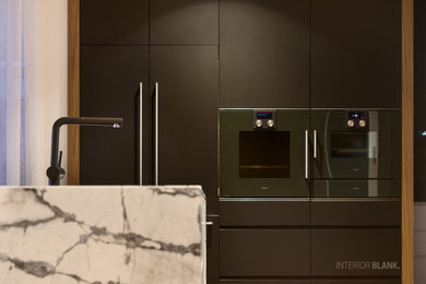 This is an example of an expansive contemporary kitchen pantry in Brisbane with an undermount sink, flat-panel cabinets, black cabinets, marble benchtops, black splashback, stone slab splashback, black appliances, medium hardwood floors, with island, brown floor and multi-coloured benchtop.