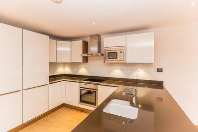 Medium sized contemporary u-shaped open plan kitchen in Channel Islands with a submerged sink, flat-panel cabinets, white cabinets, black splashback, stainless steel appliances, no island and black worktops.