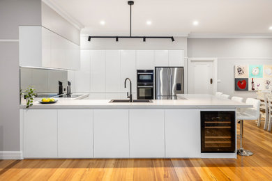 This is an example of a large modern u-shaped open plan kitchen in Melbourne with a double-bowl sink, flat-panel cabinets, white cabinets, quartz benchtops, grey splashback, glass sheet splashback, black appliances, medium hardwood floors, no island, brown floor and white benchtop.