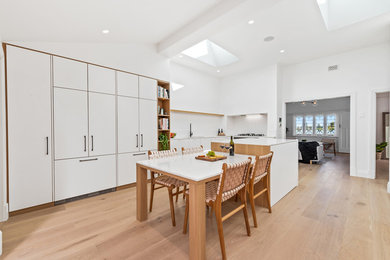 Mid-sized scandinavian l-shaped eat-in kitchen in Perth with an undermount sink, flat-panel cabinets, white cabinets, quartz benchtops, white splashback, mosaic tile splashback, black appliances, medium hardwood floors, with island, brown floor and white benchtop.