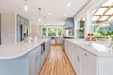 This is an example of an expansive country l-shaped kitchen pantry in Portland with a farmhouse sink, shaker cabinets, blue cabinets, quartz benchtops, white splashback, ceramic splashback, stainless steel appliances, light hardwood floors, with island, multi-coloured floor and white benchtop.