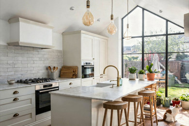 Inspiration for a medium sized modern grey and white galley kitchen in London with a belfast sink, shaker cabinets, white cabinets, marble worktops, grey splashback, porcelain splashback, stainless steel appliances, dark hardwood flooring, an island, brown floors, white worktops and a vaulted ceiling.