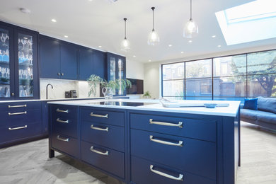 Design ideas for a large classic open plan kitchen in Berkshire with an integrated sink, beaded cabinets, blue cabinets, engineered stone countertops, white splashback, black appliances, light hardwood flooring, an island and white worktops.