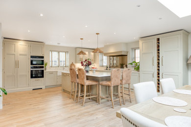 This is an example of an expansive country grey and brown l-shaped open plan kitchen in Hertfordshire with a belfast sink, shaker cabinets, beige cabinets, wood worktops, metallic splashback, mirror splashback, black appliances, light hardwood flooring, an island, beige floors, white worktops, a coffered ceiling and a feature wall.