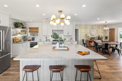 Inspiration for a large transitional l-shaped laminate floor and brown floor kitchen pantry remodel in San Diego with a farmhouse sink, shaker cabinets, white cabinets, quartz countertops, white backsplash, ceramic backsplash, stainless steel appliances, an island and multicolored countertops