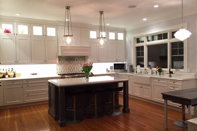 Large transitional u-shaped medium tone wood floor eat-in kitchen photo in New York with a farmhouse sink, recessed-panel cabinets, white cabinets, quartz countertops, white backsplash, an island and stainless steel appliances