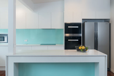 This is an example of a large beach style l-shaped eat-in kitchen in Brisbane with a drop-in sink, raised-panel cabinets, white cabinets, quartz benchtops, blue splashback, glass sheet splashback, stainless steel appliances, medium hardwood floors and with island.