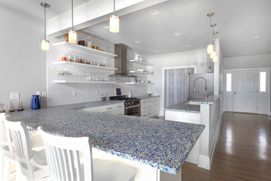 This is an example of a mid-sized beach style galley open plan kitchen in Charleston with white cabinets, a peninsula, recycled glass benchtops, white splashback, medium hardwood floors, a double-bowl sink, flat-panel cabinets, subway tile splashback, stainless steel appliances and beige floor.