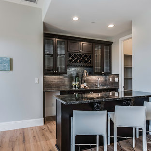 サクラメントの小さいトランジショナルスタイルのおしゃれな着席型バー (アンダーカウンターシンク、ガラス扉のキャビネット、濃色木目調キャビネット、御影石カウンター、黒いキッチンパネル、スレートの床、淡色無垢フローリング、ベージュの床) の写真