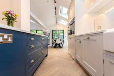 Medium sized contemporary kitchen/diner in Hertfordshire with shaker cabinets, blue cabinets, composite countertops, glass sheet splashback, stainless steel appliances, light hardwood flooring, an island, white worktops, a vaulted ceiling and feature lighting.