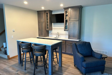 Example of a small transitional laminate floor and brown floor wet bar design in Minneapolis with an undermount sink, recessed-panel cabinets, brown cabinets, quartz countertops, beige backsplash, ceramic backsplash and white countertops