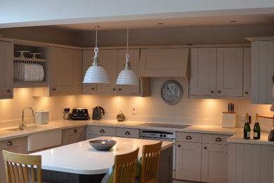 Photo of a large traditional u-shaped kitchen in London with a farmhouse sink, shaker cabinets, grey cabinets, solid surface benchtops, white splashback, black appliances, medium hardwood floors and with island.