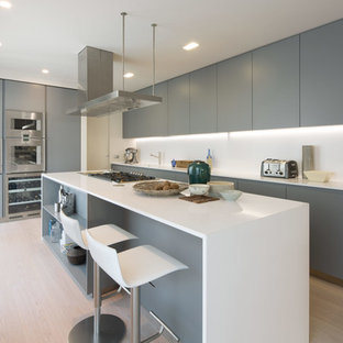 Modelo de cocina actual con armarios con paneles lisos, puertas de armario grises, salpicadero blanco, electrodomésticos de acero inoxidable, suelo de madera clara, una isla, suelo beige y encimeras blancas