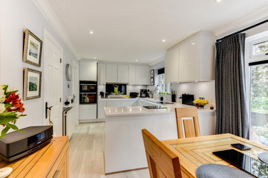 Medium sized grey and white u-shaped kitchen/diner in Sussex with a submerged sink, flat-panel cabinets, white cabinets, quartz worktops, multi-coloured splashback, glass sheet splashback, black appliances, vinyl flooring, a breakfast bar, grey floors, white worktops and feature lighting.