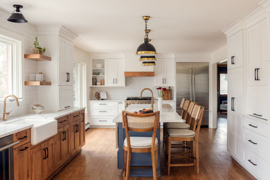 Example of a transitional u-shaped medium tone wood floor and brown floor kitchen design in Chicago with a farmhouse sink, shaker cabinets, white cabinets, white backsplash, subway tile backsplash, stainless steel appliances, an island and white countertops