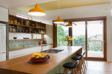 This is an example of a mid-sized contemporary single-wall open plan kitchen in Wollongong with a double-bowl sink, flat-panel cabinets, white cabinets, wood benchtops, green splashback, mosaic tile splashback, white appliances, light hardwood floors and with island.