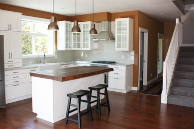 This is an example of a mid-sized arts and crafts l-shaped eat-in kitchen in Vancouver with a farmhouse sink, shaker cabinets, white cabinets, granite benchtops, grey splashback, subway tile splashback, stainless steel appliances, medium hardwood floors, with island and brown floor.