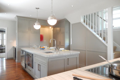 Photo of a traditional u-shaped kitchen in Philadelphia with a farmhouse sink, shaker cabinets, grey cabinets, quartz benchtops, grey splashback, stainless steel appliances, medium hardwood floors, with island, brown floor and grey benchtop.