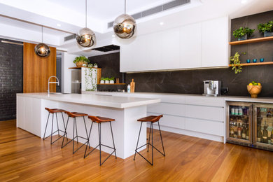 Inspiration for an expansive contemporary galley open plan kitchen in Brisbane with a drop-in sink, flat-panel cabinets, quartz benchtops, black splashback, marble splashback, stainless steel appliances, medium hardwood floors, with island, brown floor and white cabinets.