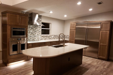 Example of a mid-sized transitional l-shaped medium tone wood floor and brown floor enclosed kitchen design in Jacksonville with a farmhouse sink, shaker cabinets, medium tone wood cabinets, granite countertops, multicolored backsplash, porcelain backsplash, stainless steel appliances, an island and beige countertops