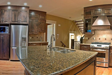 Mid-sized arts and crafts l-shaped light wood floor and brown floor open concept kitchen photo in Calgary with a double-bowl sink, raised-panel cabinets, dark wood cabinets, granite countertops, beige backsplash, stone tile backsplash, stainless steel appliances and an island