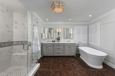 This is an example of a traditional bathroom in DC Metro with grey cabinets, a freestanding tub, a corner shower, white tile, marble, grey walls, wood-look tile, an undermount sink, engineered quartz benchtops, brown floor, a hinged shower door, white benchtops, a shower seat, a double vanity, raised-panel cabinets, a built-in vanity and panelled walls.