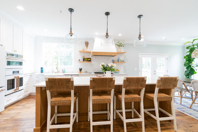 Photo of a mid-sized country l-shaped eat-in kitchen in Baltimore with a farmhouse sink, shaker cabinets, light wood cabinets, quartz benchtops, multi-coloured splashback, ceramic splashback, white appliances, light hardwood floors, with island, brown floor and multi-coloured benchtop.