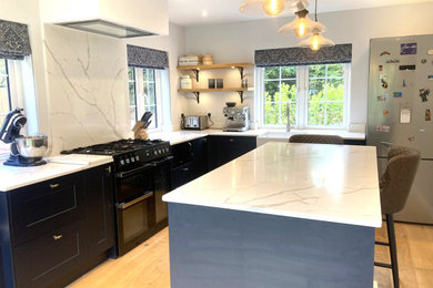 This is an example of a medium sized classic kitchen/diner in Surrey with a belfast sink, shaker cabinets, blue cabinets, quartz worktops, white splashback, marble splashback, black appliances, light hardwood flooring, an island, white worktops and feature lighting.