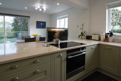 Photo of a modern kitchen in Gloucestershire with a belfast sink, shaker cabinets, green cabinets, engineered stone countertops, beige splashback, an island, black floors and beige worktops.