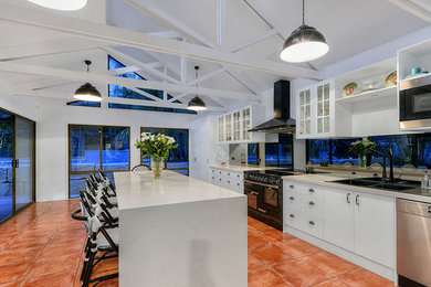 This is an example of a large contemporary galley open plan kitchen in Brisbane with a drop-in sink, glass-front cabinets, white cabinets, solid surface benchtops, grey splashback, glass sheet splashback, black appliances, terra-cotta floors and with island.