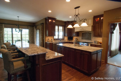 Inspiration for a large timeless galley medium tone wood floor eat-in kitchen remodel in Milwaukee with an undermount sink, raised-panel cabinets, dark wood cabinets, granite countertops, beige backsplash, stone tile backsplash, stainless steel appliances and two islands