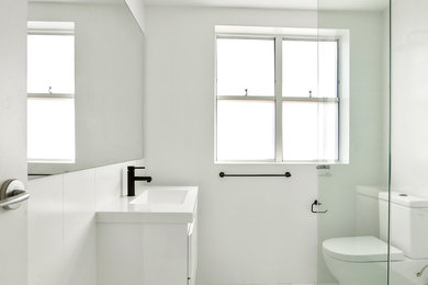 Inredning av ett modernt badrum, med släta luckor, vita skåp, en toalettstol med hel cisternkåpa, vit kakel, porslinskakel, vita väggar, klinkergolv i porslin, ett integrerad handfat och vitt golv