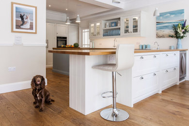 Mid-sized country u-shaped eat-in kitchen in Kent with a single-bowl sink, beaded inset cabinets, white cabinets, wood benchtops, white splashback, black appliances, medium hardwood floors, with island, brown floor and brown benchtop.