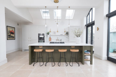 This is an example of a large classic single-wall open plan kitchen in Cardiff with a belfast sink, shaker cabinets, light wood cabinets, quartz worktops, white splashback, stone slab splashback, stainless steel appliances, porcelain flooring, an island, white worktops, a vaulted ceiling and feature lighting.